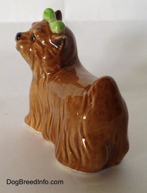 The back left side of a figurine of a brown Yorkshire Terrier with a bow in its hair. The figurine has a short tail that is in the air.